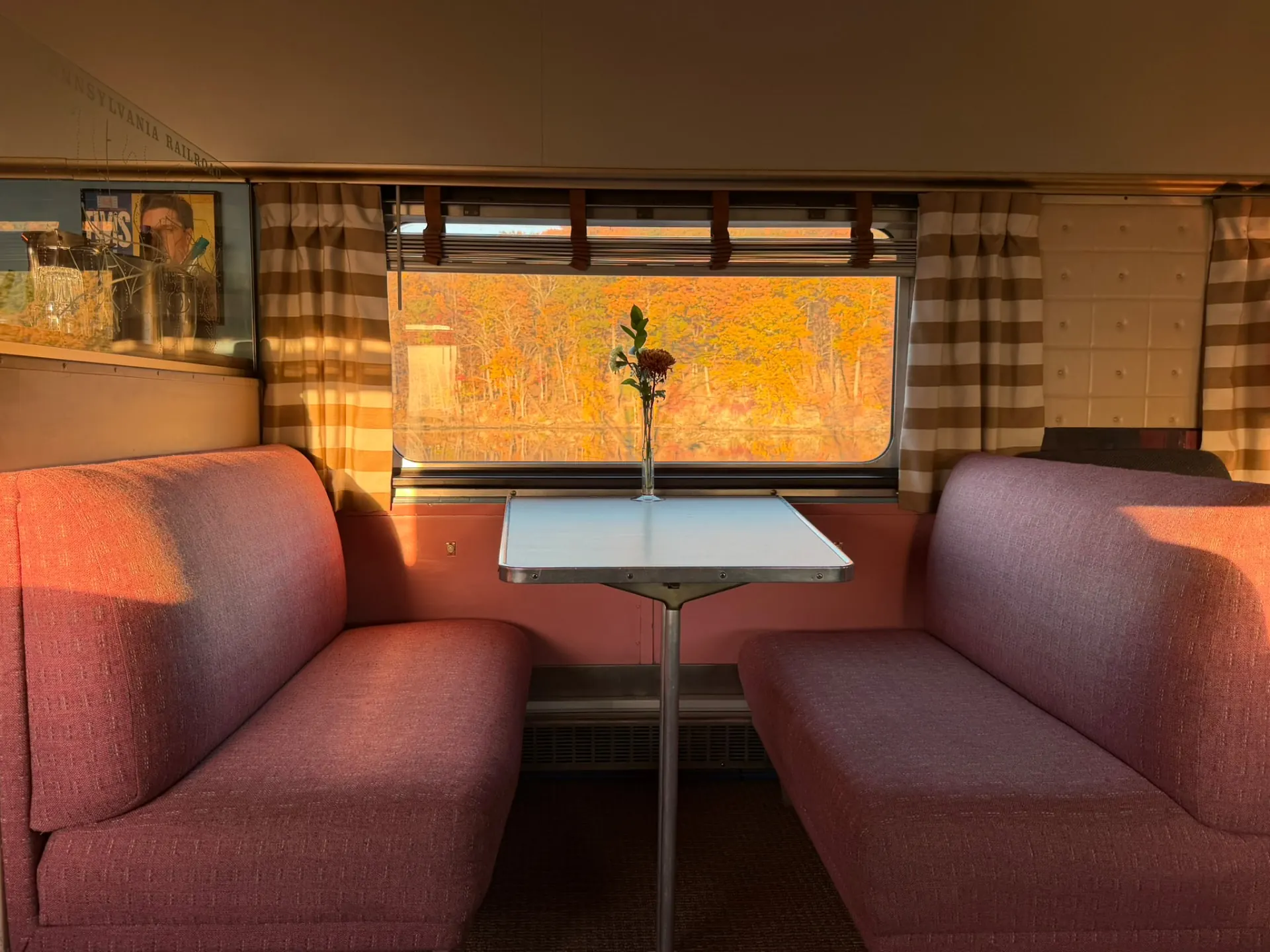 Pink booth seats and a table with a single flower inside a 1949 Pullman car, autumn trees outside the window